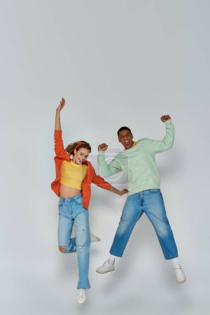happy interracial couple in casual attire jumping together on grey backdrop, youthful spirit
