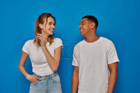 Photo for Cheerful woman posing with hand in pocket and looking at african american friend on blue background - Royalty Free Image