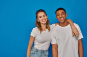 happy woman hugging african american man and looking at camera on blue backdrop, cultural diversity Stickers #671418106