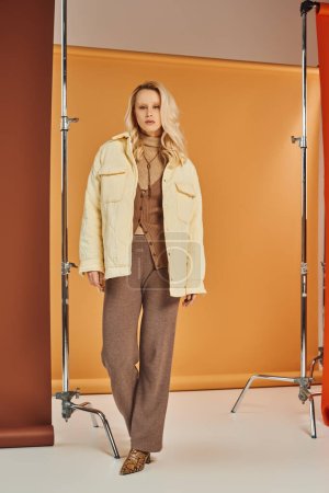 Blondine in stylischer Oberbekleidung und Stiefeln blickt in die Kamera und posiert im Atelier, Herbstmode