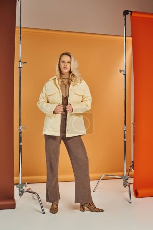 Blondes Model in stylischer Oberbekleidung und Stiefeln blickt in die Kamera, posiert im Studio, Herbstmode