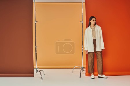 stylish asian woman in autumn attire standing on multicolored backdrop, leather pants and outerwear