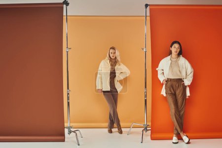 Herbstmode, junge interrassische Frauen in stilvoller Kleidung posieren vor pastellfarbener Kulisse