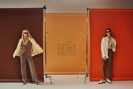 guardarropa de otoño, elegantes modelos multiétnicos en gafas de sol y ropa de abrigo posando sobre colorido telón de fondo