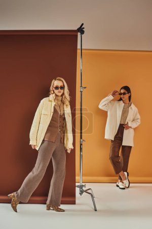 fall wardrobe, stylish multiethnic women in sunglasses and outerwear posing on duo color backdrop