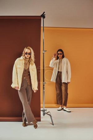 fall season, multiethnic women in sunglasses and autumn outerwear posing on duo color backdrop