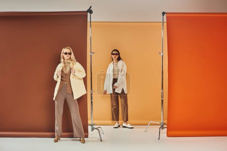 fall wardrobe, multiethnic women in sunglasses and autumn outerwear posing on colorful backdrop