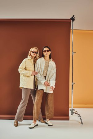 Herbstsaison, interrassische Frauen in Sonnenbrille und Oberbekleidung posieren gemeinsam auf Duo-Farbkulisse