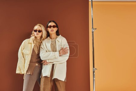 fall fashion and trends, interracial women in sunglasses and outerwear posing on duo color backdrop