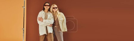 fall trends, interracial women in sunglasses and outerwear posing on duo color backdrop, banner