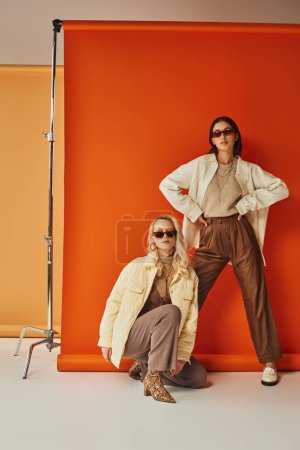 fashion and trends, interracial women in sunglasses and outerwear posing in studio, fall colors