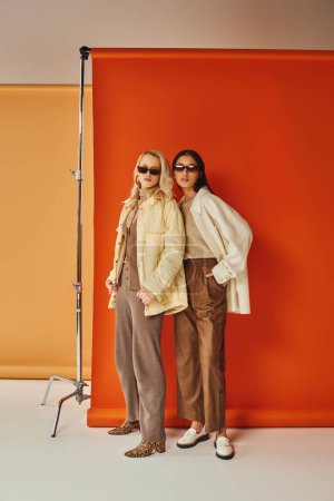 multiethnic models in sunglasses and autumn outerwear posing in studio, fall colors, duo backdrop
