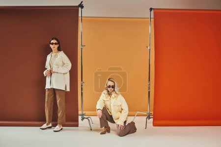 interracial models in sunglasses and autumn outerwear posing on colorful backdrop, fall colors