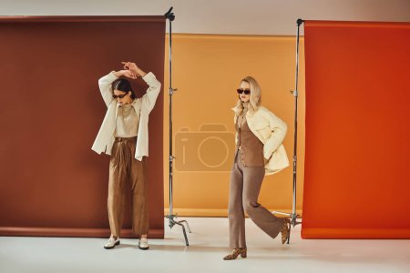 diverse cultures women in outerwear and sunglasses posing on colorful backdrop, fall fashion