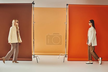 Foto de Modelos interracial en ropa de abrigo y gafas de sol caminando cerca de telón de fondo colorido, concepto de moda de otoño - Imagen libre de derechos