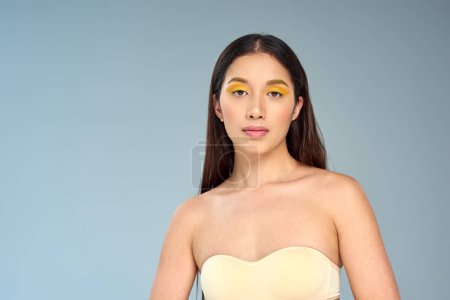 asian model with bold makeup posing in strapless top isolated on blue, diverse beauty and eye makeup