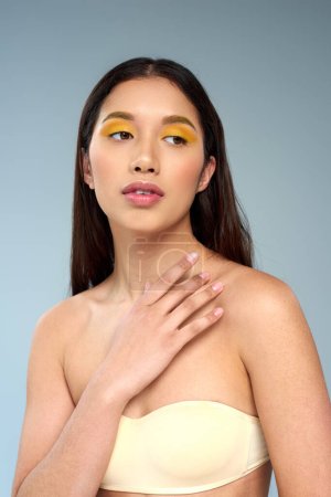 young asian model with bold makeup posing in strapless top isolated on blue, diverse beauty concept