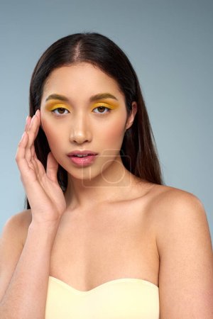 junge Asiatin mit kühnem Make-up posiert in trägerlosem Top isoliert auf blauer, strahlender Haut und Schönheit