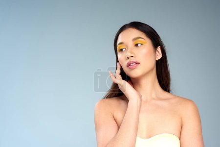 Asiatin mit kühnem Make-up posiert in trägerlosem Top isoliert auf blauer, strahlender Haut und Jugend