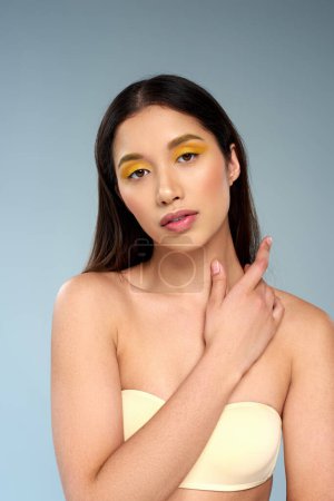 asian woman with bold makeup posing in strapless bra isolated on blue, radiant skin and youth