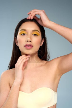 Asiatin mit kühnem Make-up posiert in trägerlosem BH isoliert auf blauer, strahlender Haut und Visage
