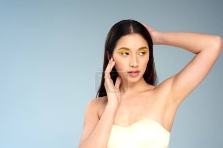 mujer asiática con maquillaje audaz posando en sujetador sin tirantes sobre fondo azul, piel radiante y rostro
