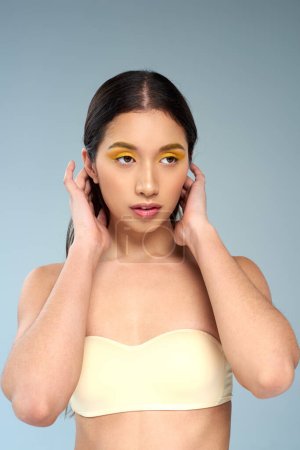 asiatisches Model mit fettem Make-up posiert in trägerlosem BH vor blauem Hintergrund, strahlende Haut