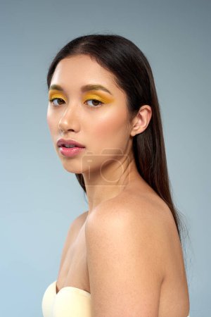 schönes Model mit fettem Make-up und nackten Schultern posiert auf blauem Hintergrund, asiatisches Schönheitskonzept