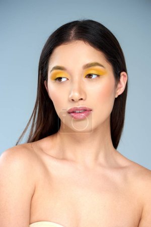 weibliche Sinnlichkeit, junges asiatisches Model mit kühnem Make-up und nackten Schultern posiert vor blauem Hintergrund