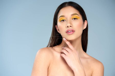 elegance, asian woman with bold makeup and bare shoulders touching chin and posing on blue backdrop
