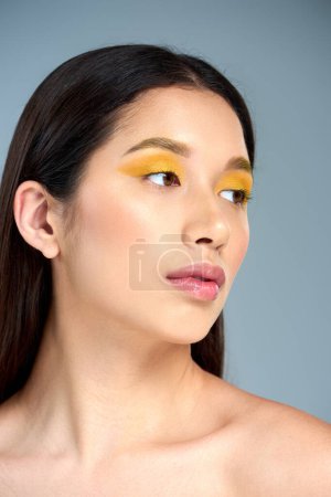 beauty and youth, asian woman with bold makeup and bare shoulders looking away on blue backdrop