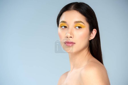 portrait, asian woman with bold makeup and bare shoulders looking away on blue backdrop, tenderness