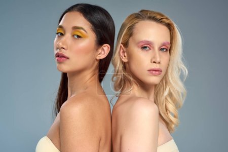 interracial models with vibrant eye makeup looking at camera on blue backdrop, beauty trend concept