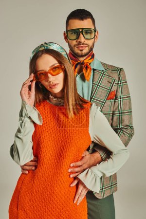 Mann im karierten Blazer umarmt Frau in orangefarbenem Kleid und Sonnenbrille auf grauer, retro-inspirierter Mode