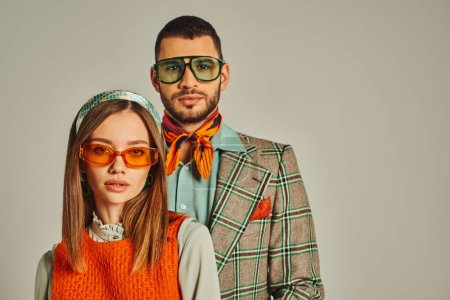 young couple in elegant vintage clothes and sunglasses looking at camera on grey, fashion from past