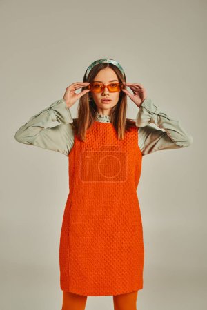 young woman in orange dress and headband adjusting sunglasses on grey, retro-inspired fashion