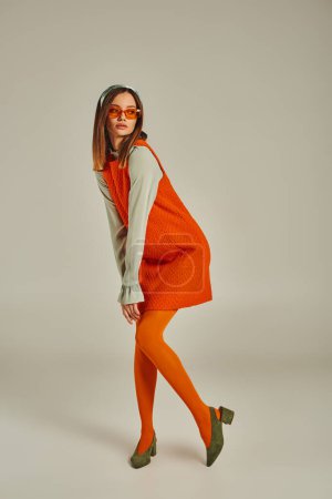 Frau im Vintage-Stil in orangefarbenem Kleid, Strumpfhose und Sonnenbrille, die auf grau wegschaut