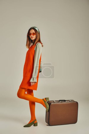 mujer de moda en vestido naranja y gafas de sol posando cerca de la maleta vintage en gris, estilo retro