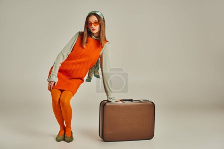 Glamour-Frau in orangefarbenem Kleid posiert in der Nähe von Vintage-Koffer auf grau, Mode aus der Vergangenheit