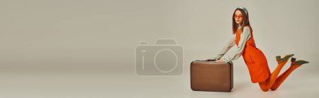 young woman in stylish retro outfit and sunglasses kneeling near vintage suitcase on grey, banner