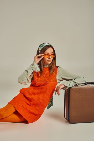 stylish woman in orange dress and sunglasses sitting near vintage suitcase and looking away on grey