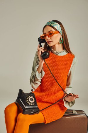 Retro-inspirierte Frau mit Sonnenbrille spricht am Wählscheibentelefon, während sie auf einem Vintage-Koffer auf grau sitzt