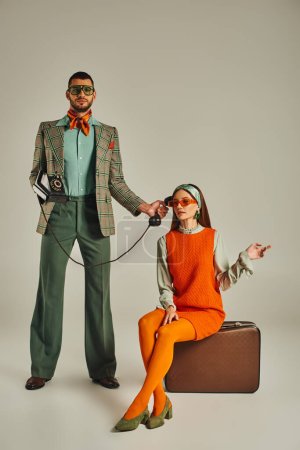 trendy man holding rotary phone near woman sitting on vintage suitcase on grey, retro lifestyle