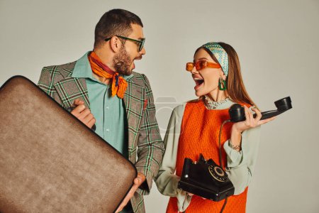 pareja de estilo retro lleno de alegría con maleta vintage y teléfono con cable mirándose el uno al otro en gris