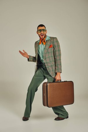 Überglücklicher Mann in kariertem Blazer und Sonnenbrille posiert mit Vintage-Koffer auf grauem Retro-Stil