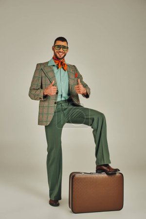 glücklicher Mann in Retro-Klamotten und Sonnenbrille zeigt Daumen hoch neben Vintage-Koffer auf grau