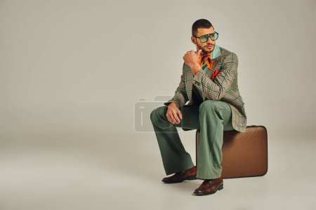 Nachdenklicher Mann im Vintage-Stil mit Sonnenbrille sitzt auf einem Retro-Koffer und schaut weg auf grau