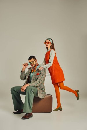 mujer en vestido naranja cerca de hombre elegante sentado en la maleta de la vendimia en gris, viajeros anticuados