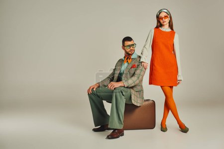 trendy man sitting on vintage suitcase near woman in orange dress on grey, retro-inspired couple