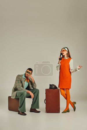 Frau in orangefarbenem Kleid spricht am Schnurtelefon neben gelangweiltem Mann, der auf einem Vintage-Koffer auf grau sitzt